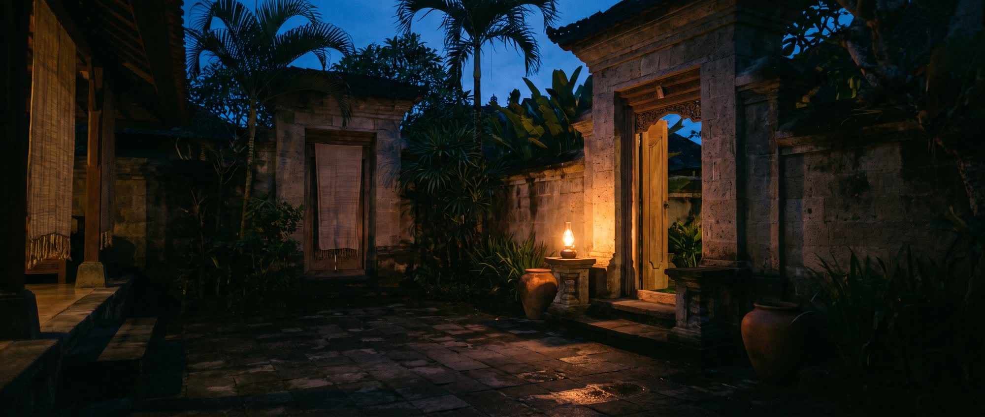 The courtyard of a Balinese compound at dusk, one warm lamp lit beside an open doorway beneath a deep blue sky.