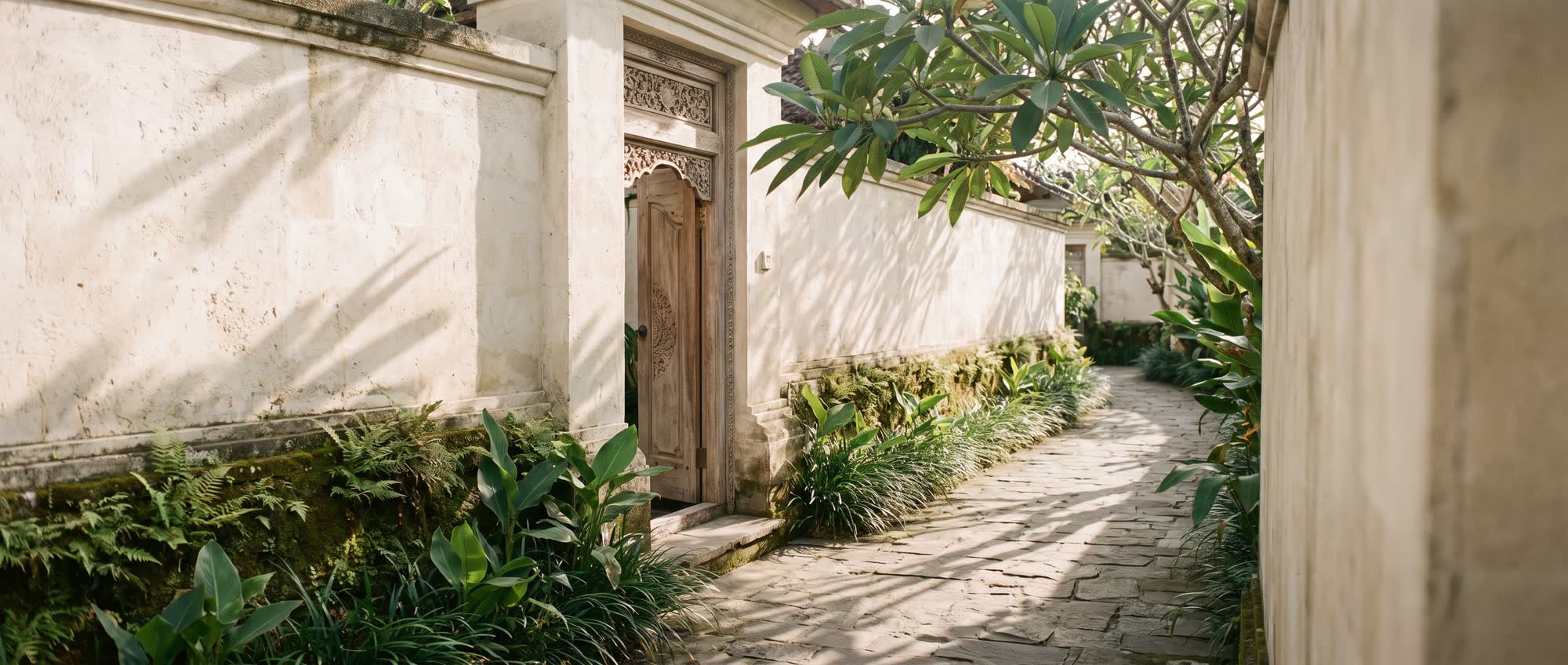 Morning light falling across the paved walkway of a Balinese compound, past a carved teak door and mossy planting beds.