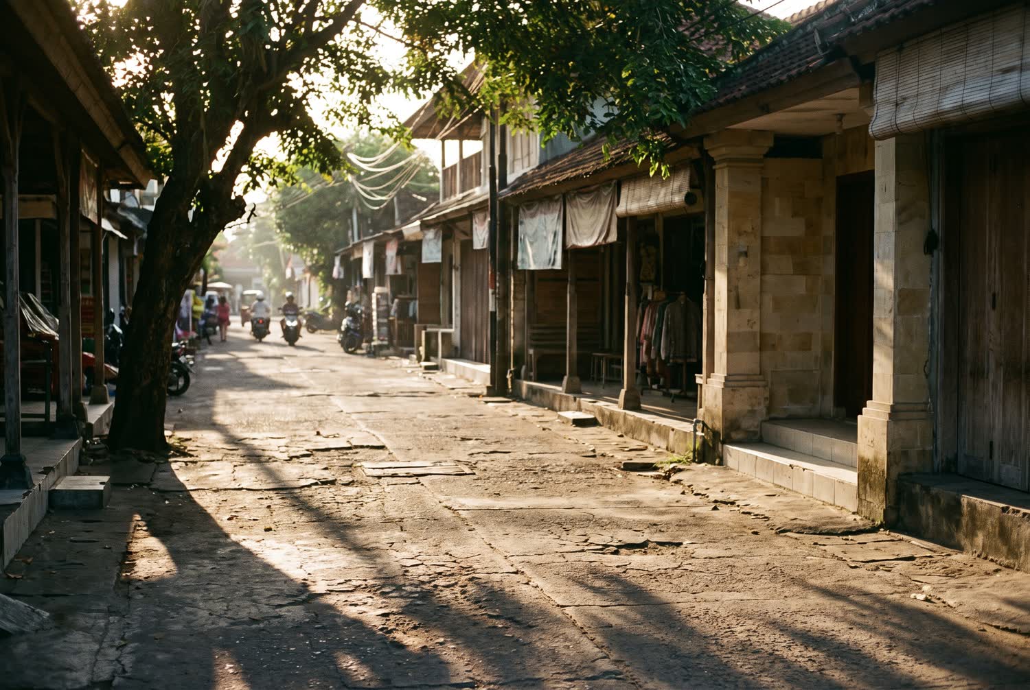 An ordinary lively street in Kuta in late afternoon light with low shopfronts and tree shadow across the road.