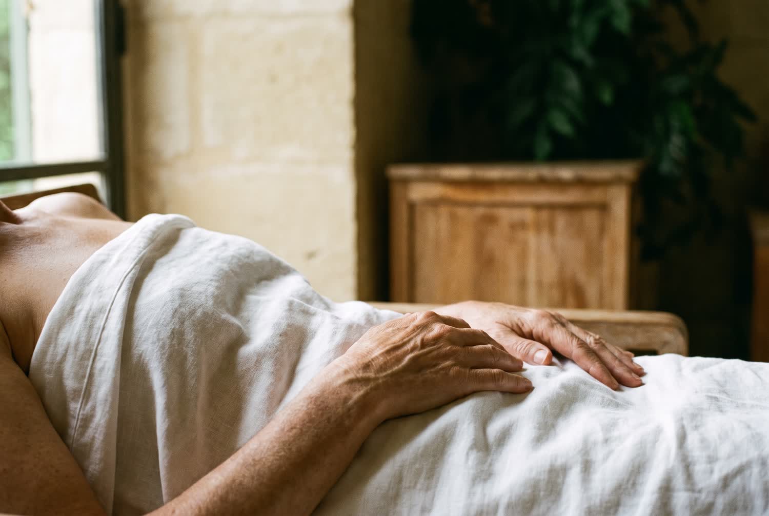 Two hands resting calmly on a shoulder covered by a white linen sheet in soft window light.