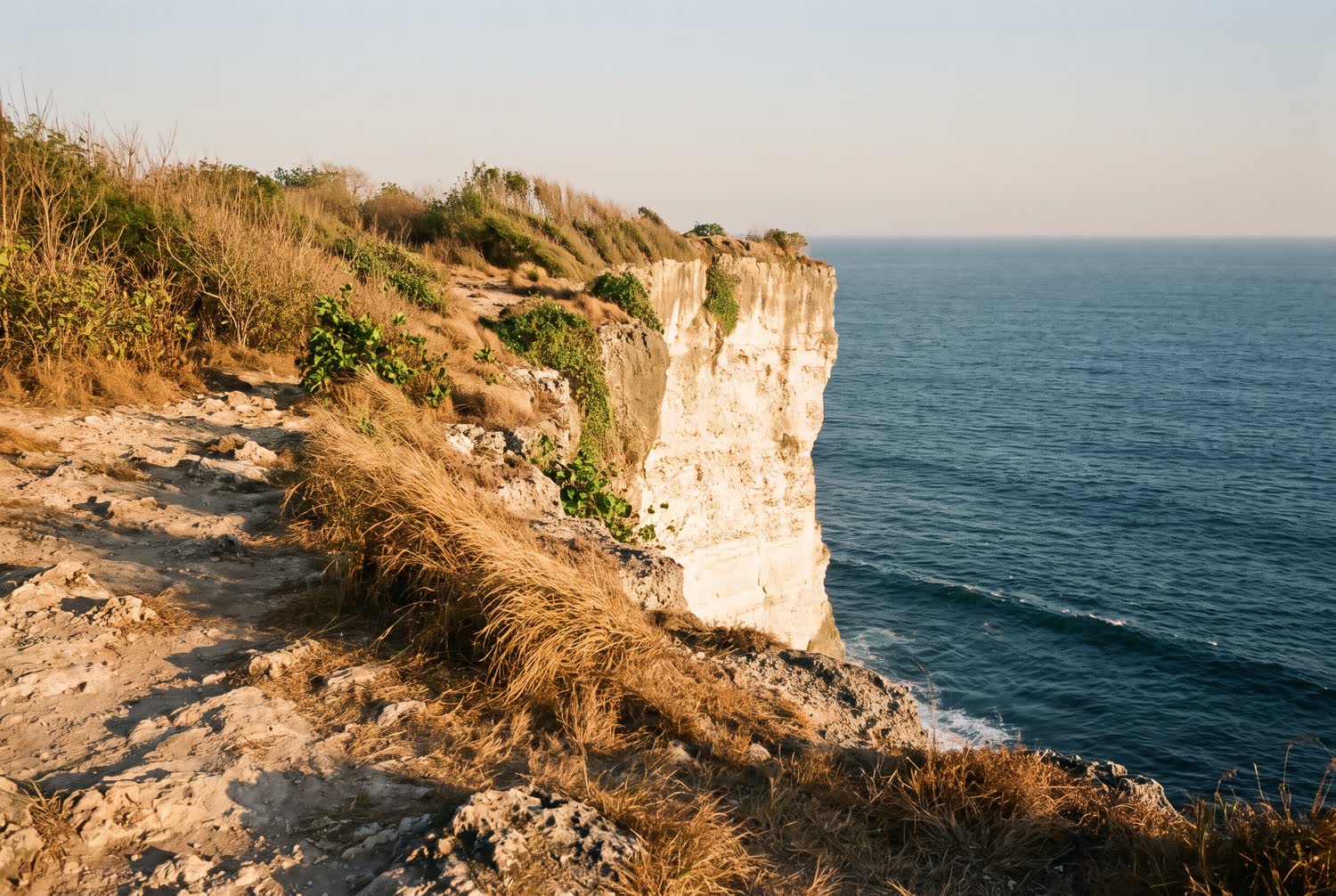 Pale limestone cliffs and dry coastal scrub in Uluwatu, with long late shadows falling towards the sea.